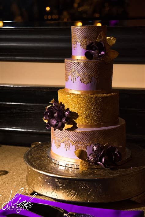 A Three Tiered Wedding Cake With Purple Flowers On The Top And Gold