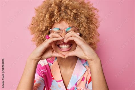 Happy Curly Haired Young Woman Makes Heart Gesture Over Mouth Smiles