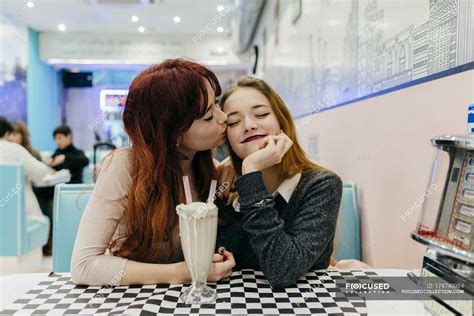 Portrait Of Redhead Girl Kissing Girlfriends Cheek At Cafe Table With