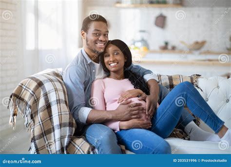 Caring Black Man Embracing His Pregnant Wife On Sofa At Home Stock