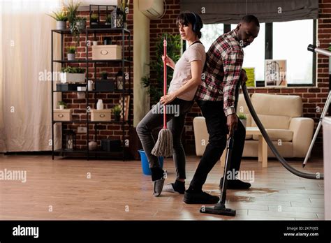 Multiracial Couple Having Fun While Cleaning The House Dancing And Singing Together Doing