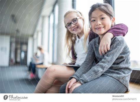 Portrait Of Two Smiling Schoolgirls Sitting On School Corridor A