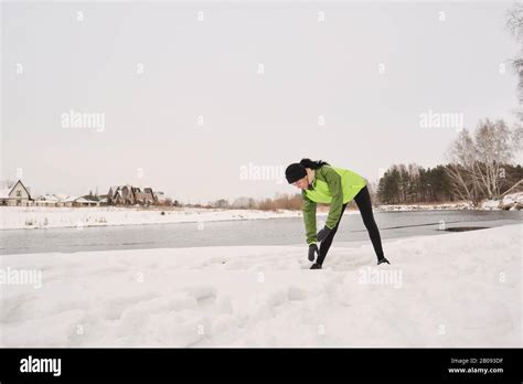 Young Woman In Green Jacket Standing T Winter Lake And Bending Over