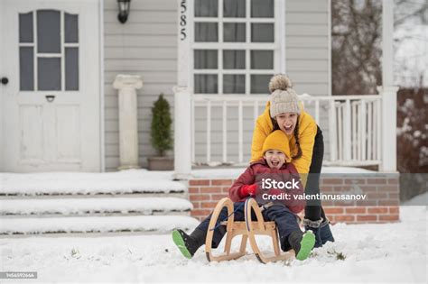 A Bigger Sister Rides Her Brother On A Sled In Front Of Their House In A Snowy Winter Frosty Day