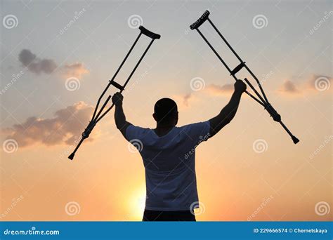 Man Raising Hands With Underarm Crutches Up To Sky Outdoors At Sunset