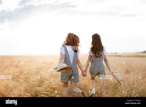 Rear View Of Happy Twin Sisters Holding Hands While Walking On Grassy Field Against Sky During