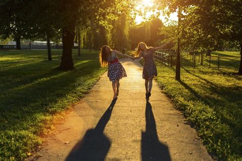 Two Young Girls Girlfriends Go Together Along The Park In Backlight Rays Of The Setting Sun