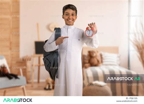 طالب عربي خليجي سعودي يرتدي الزي المدرسي ويشير باصبع يده السبابة نحو ساعة تنبيه صغيرة، حقيبة
