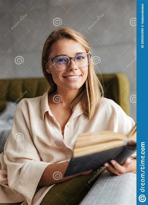 Image Of Young Smiling Woman Reading Book While Sitting On Couch Stock