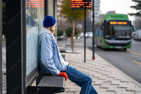 Millennial Guy Sitting On Bench At Bus Stop Looking At Arriving Bus