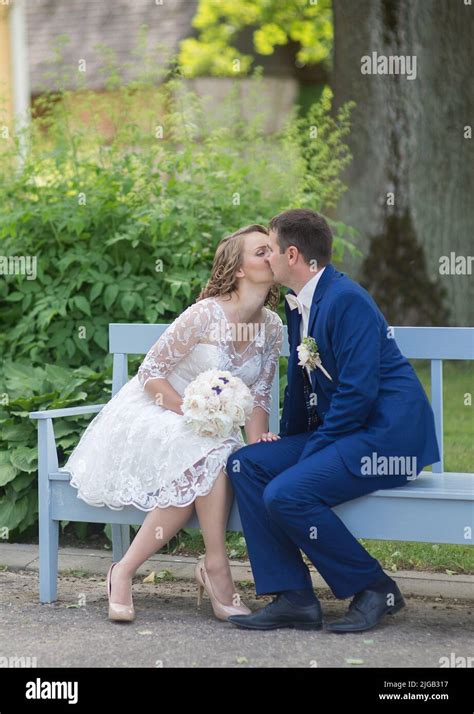 A Vertical Shot Of Newlyweds Sitting On A Bench In The Park And Kissing