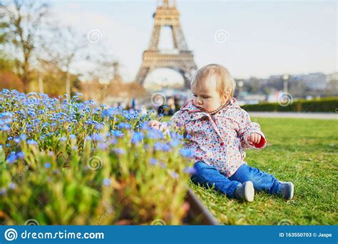 One Year Old Girl Sitting On The Grass With Blue Flowers And Eiffel