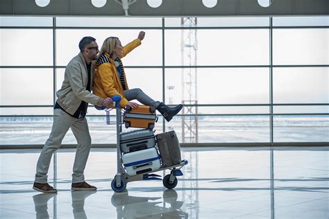 Couple On Luggage Cart In Airport Cyrus Travel Agency