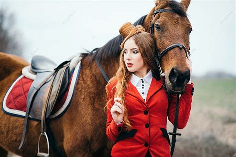 فتاة الفارس ذات الشعر الأحمر ترتدي كارديجانًا أحمر وأحذية عالية سوداء مع حصان في نزهة على