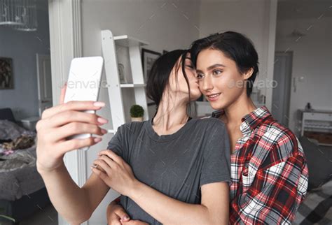 Young Happy Lesbian Couple Holding Smartphone Taking Selfie At Home