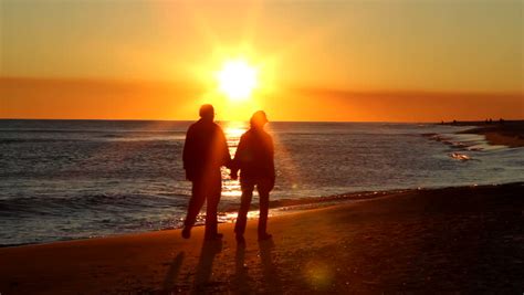Senior Adult Couple Holding Hands Walk Up The Beach As The Sun Is About