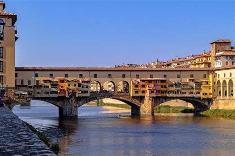 Image of Ponte Vecchio, the iconic bridge lined with shops, with the Arno River flowing below. Tourists strolling across the bridge and enjoying the scenic views.  